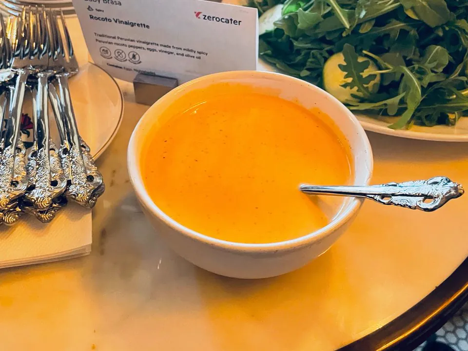 Baby Brasa rocoto vinaigrette at a client dinner A white bowl of orange rocoto vinaigrette with a Baby Brasa label card, beside an arugula salad and silver forks