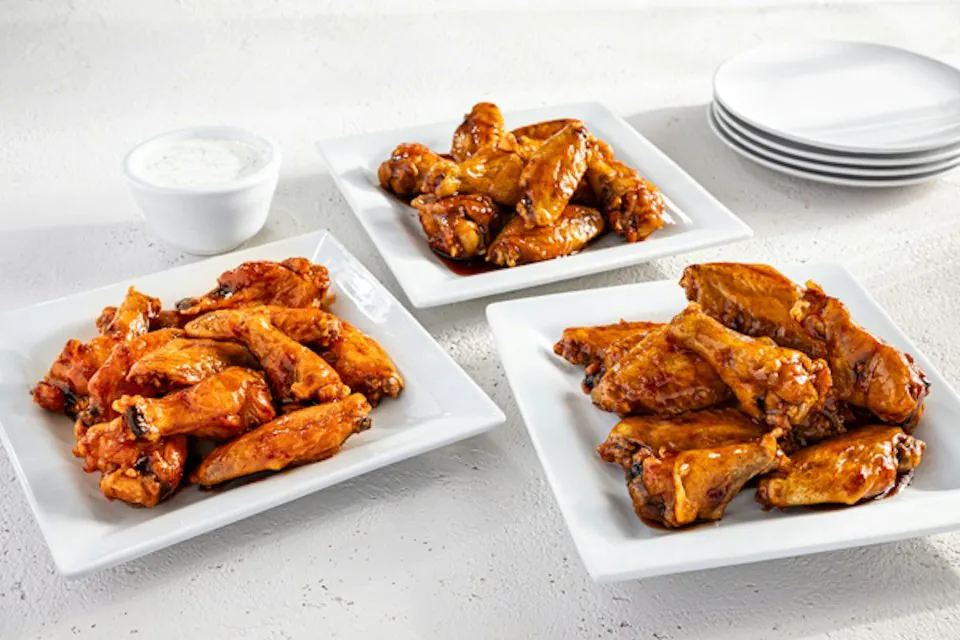 Three white plates of Chili's party platter wings in Buffalo, Honey-Chipotle and House BBQ sauce beside a bowl of ranch and a stack of plates