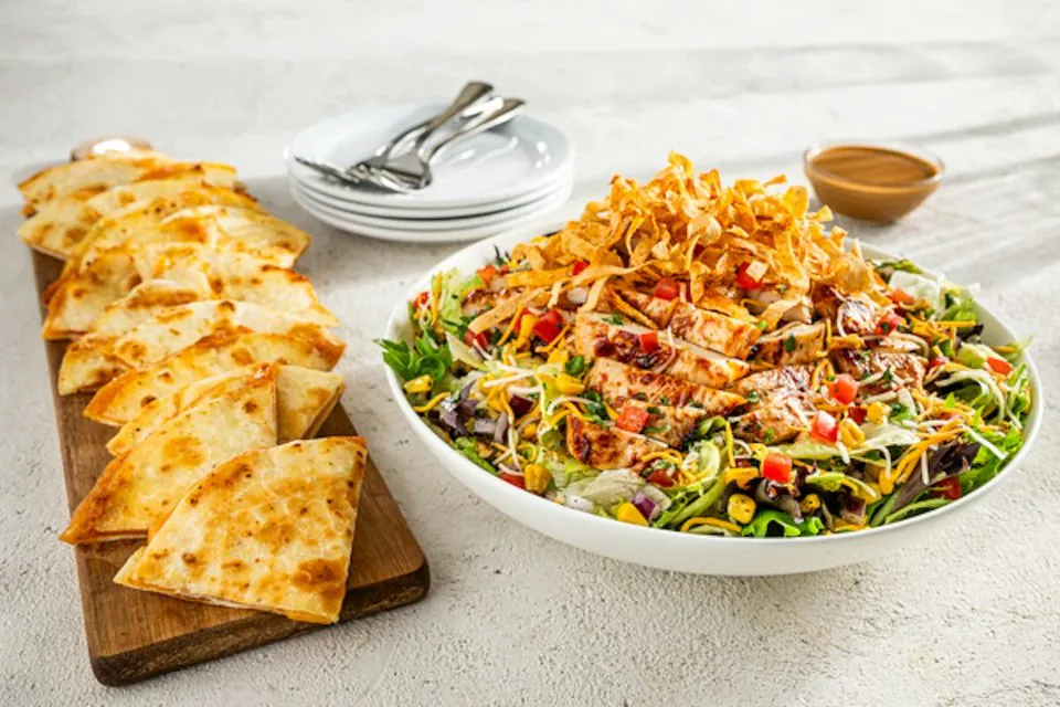 A Chili's Quesadilla Explosion Salad platter in a white bowl, topped with tortilla strips, corn salsa and tomatoes, beside a board of cheese quesadilla wedges and a cup of dressing