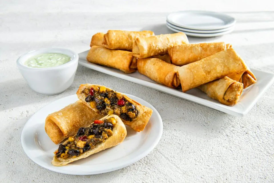 A platter of Chili's Southwestern Eggrolls with two cut open to show black beans, corn and red peppers, beside a bowl of avocado-ranch