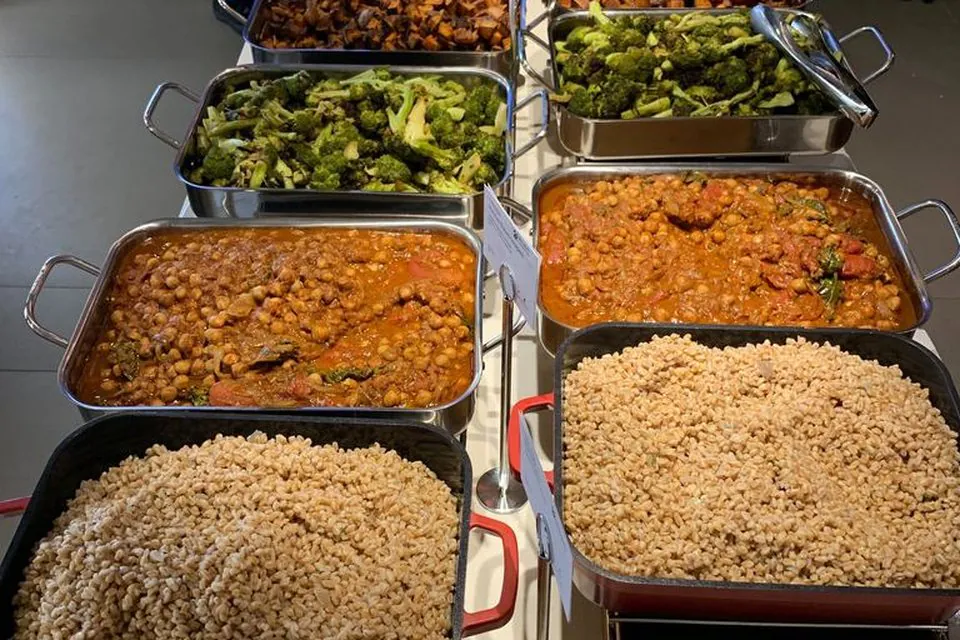 Office buffet with steel pans of roasted broccoli and roasted sweet potatoes behind pans of braised chickpeas and two trays of farro