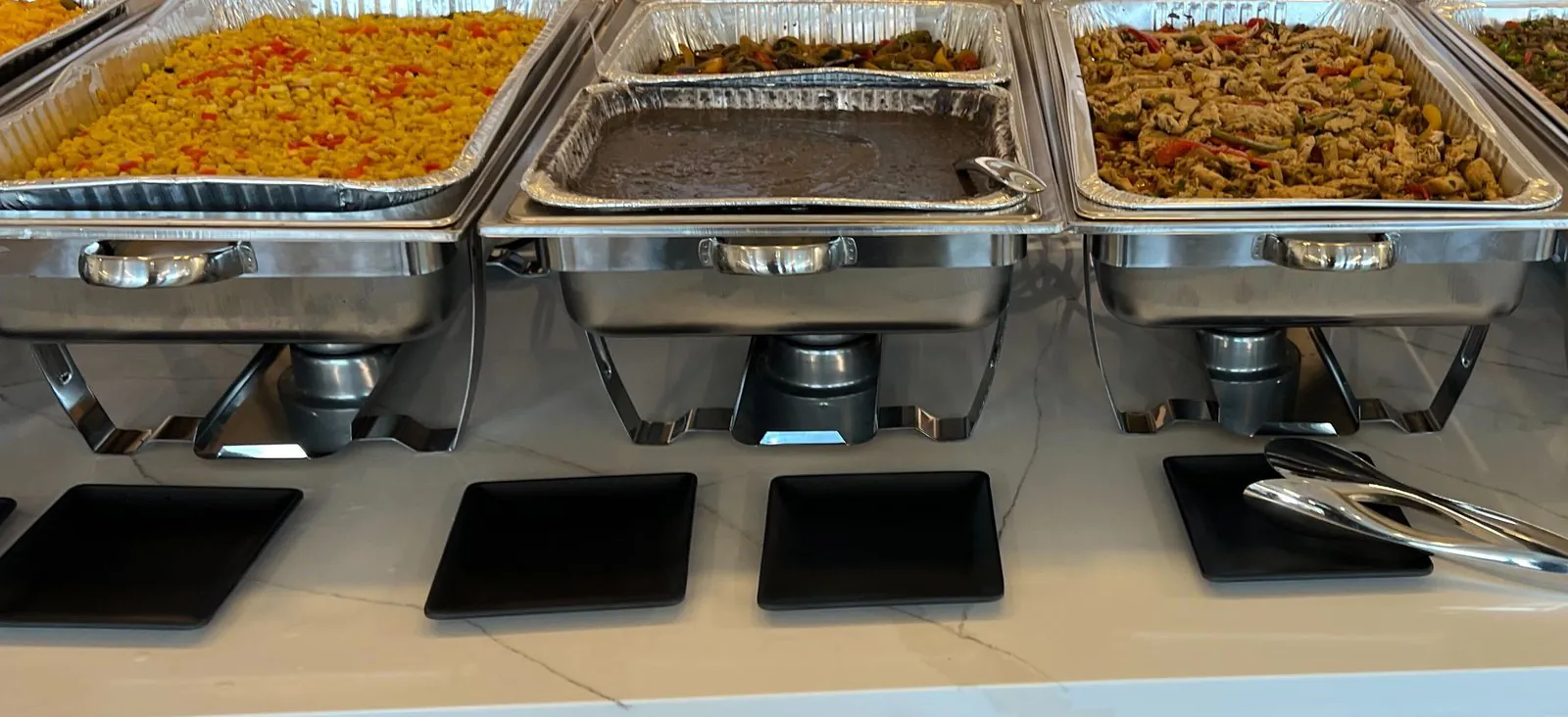 Office buffet line of chafing dishes holding roasted corn, black beans, eggplant fajitas and chicken fajitas, with black square plates on a marble counter