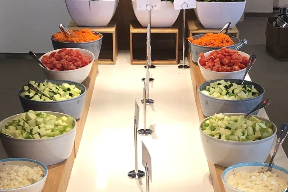 Two rows of salad bar bowls holding shredded carrots, diced tomatoes, cucumber and zucchini on a bamboo runner, with tubs of mixed greens behind
