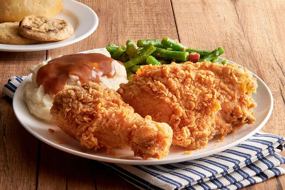 A white plate of fried chicken with mashed potatoes under brown gravy and green beans, with a yeast roll and chocolate chip cookies on a plate behind