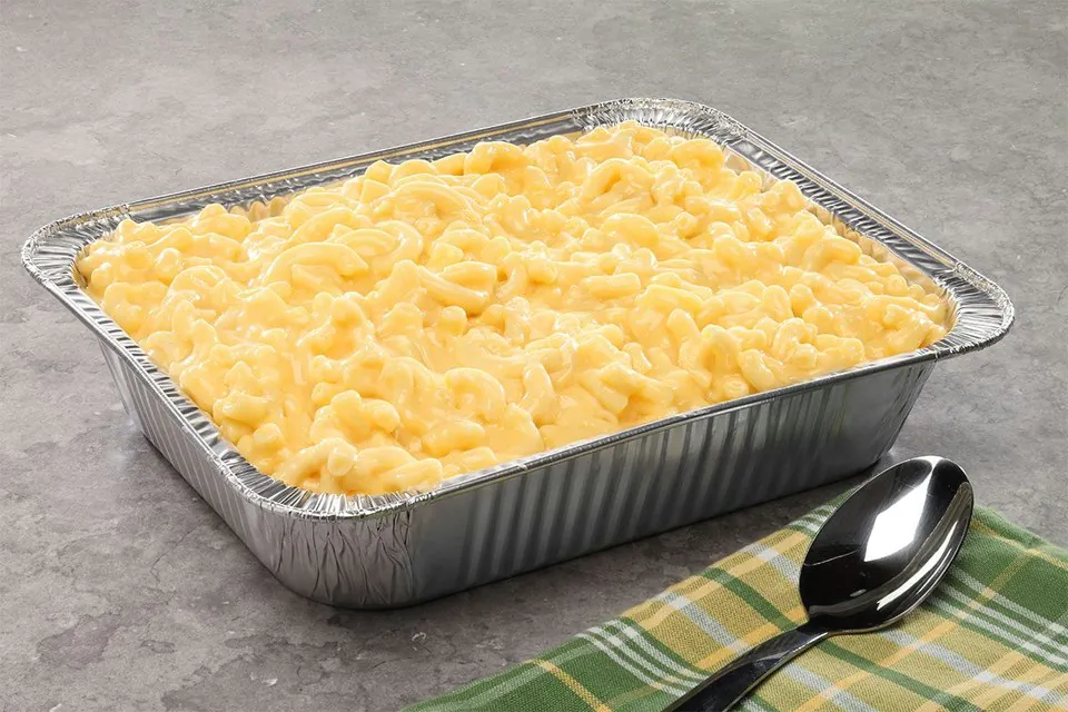 A foil catering tray of baked macaroni and cheese with a golden top, beside a serving spoon on a plaid napkin
