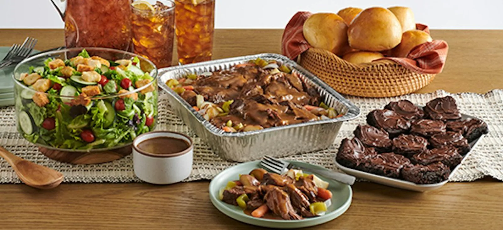 A foil tray of pot roast in gravy with carrots and onions, beside a glass bowl of garden salad, a basket of yeast rolls, a plate of brownies and a pitcher of iced tea