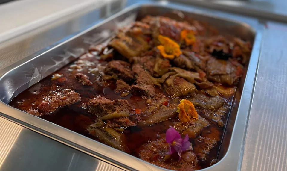 A stainless tray of braised beef and eggplant in a deep red tomato sauce, garnished with orange and pink edible flowers
