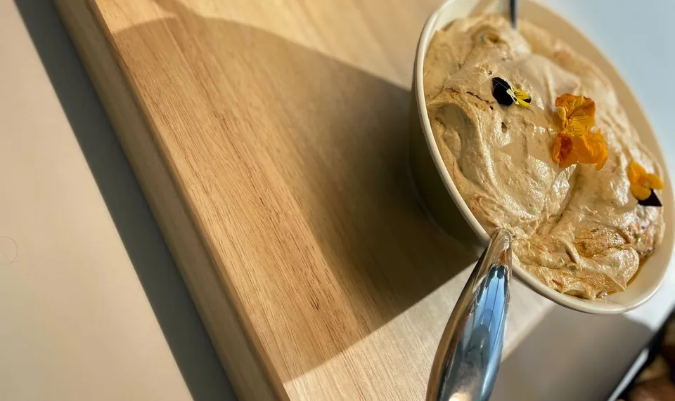 A bowl of smooth hummus garnished with orange and purple edible flowers, with a serving spoon, on a wooden board in sunlight