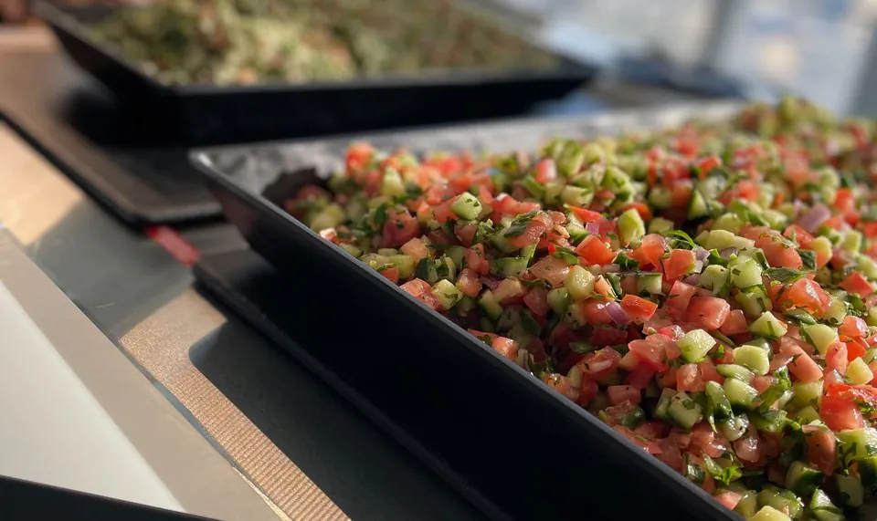 Two black platters on a buffet, a chopped tomato and cucumber salad in front and a green herb salad behind