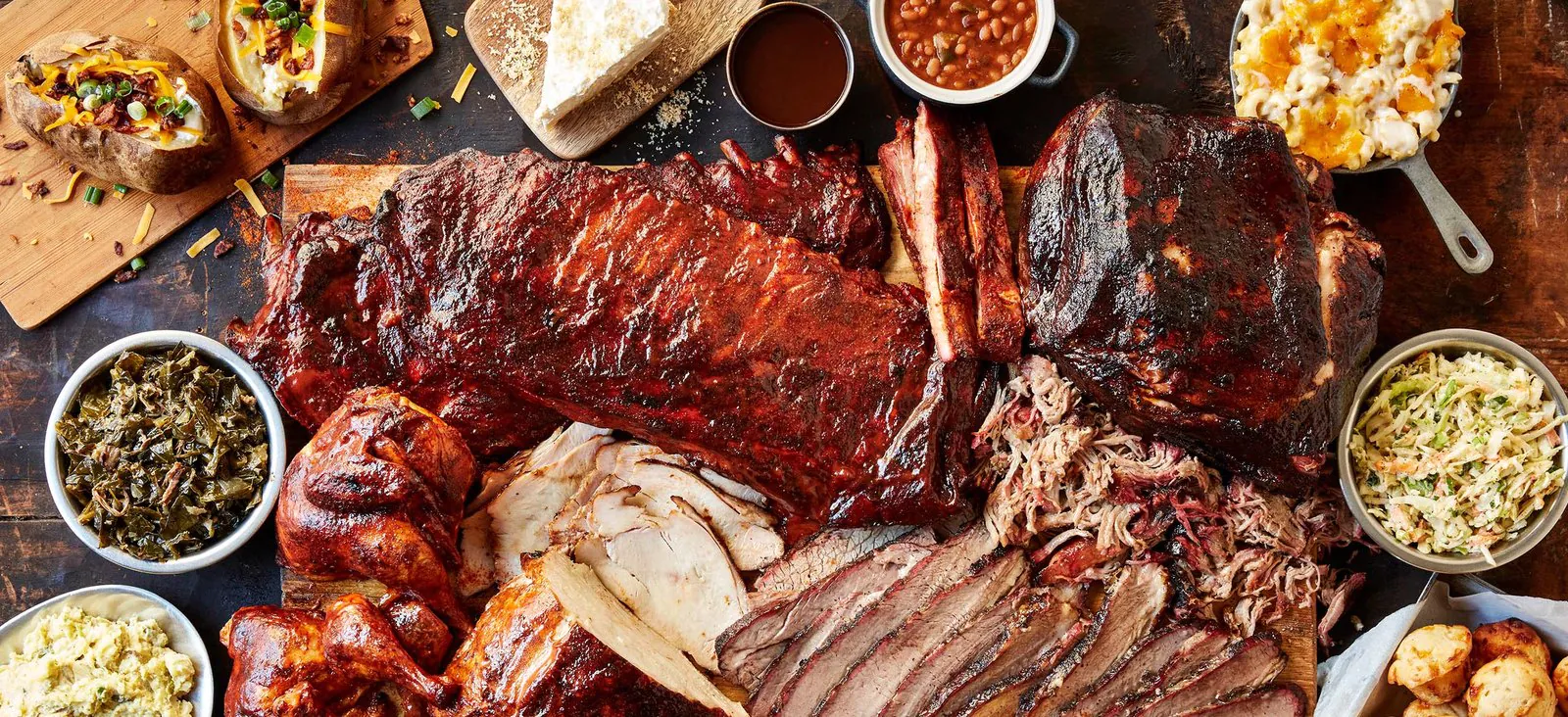 An overhead Jim 'N Nick's barbecue spread on a dark wood table: racks of spare ribs, a whole brisket, sliced turkey, pulled pork, barbecue chicken, collard greens, potato salad, baked beans, coleslaw, mac and cheese and a pile of cheese biscuits