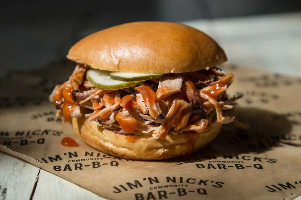 A Jim 'N Nick's pulled pork sandwich on a potato bun with pickle slices and Bar-B-Q sauce, on branded Jim 'N Nick's Community Bar-B-Q paper