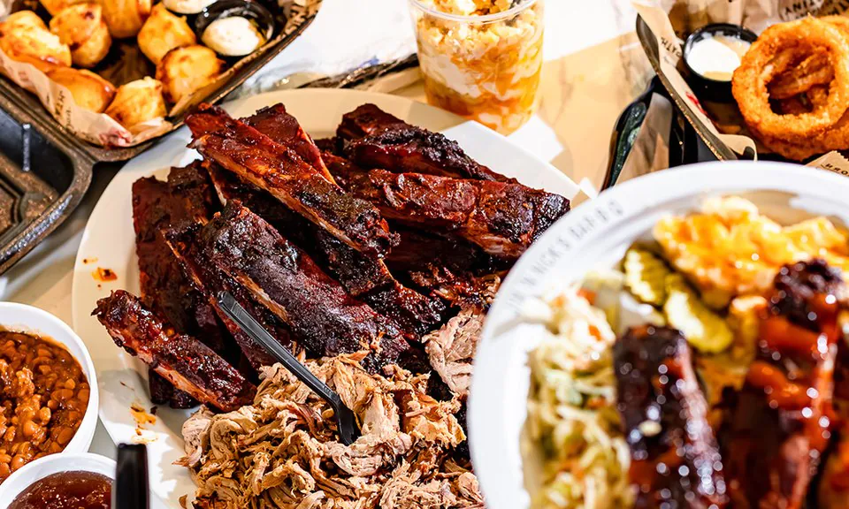 A Jim 'N Nick's table with a platter of spare ribs and pulled pork, cups of baked beans and sauce, a tray of cheese biscuits, a cup of mac and cheese and a plate of coleslaw, pickles and ribs