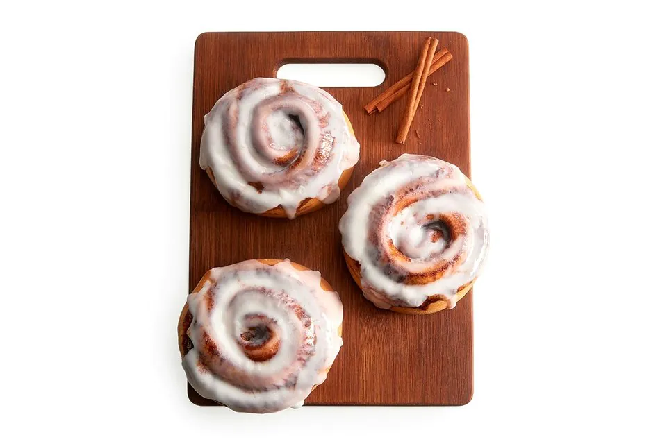 Three iced cinnamon rolls on a dark wooden cutting board with cinnamon sticks laid beside them