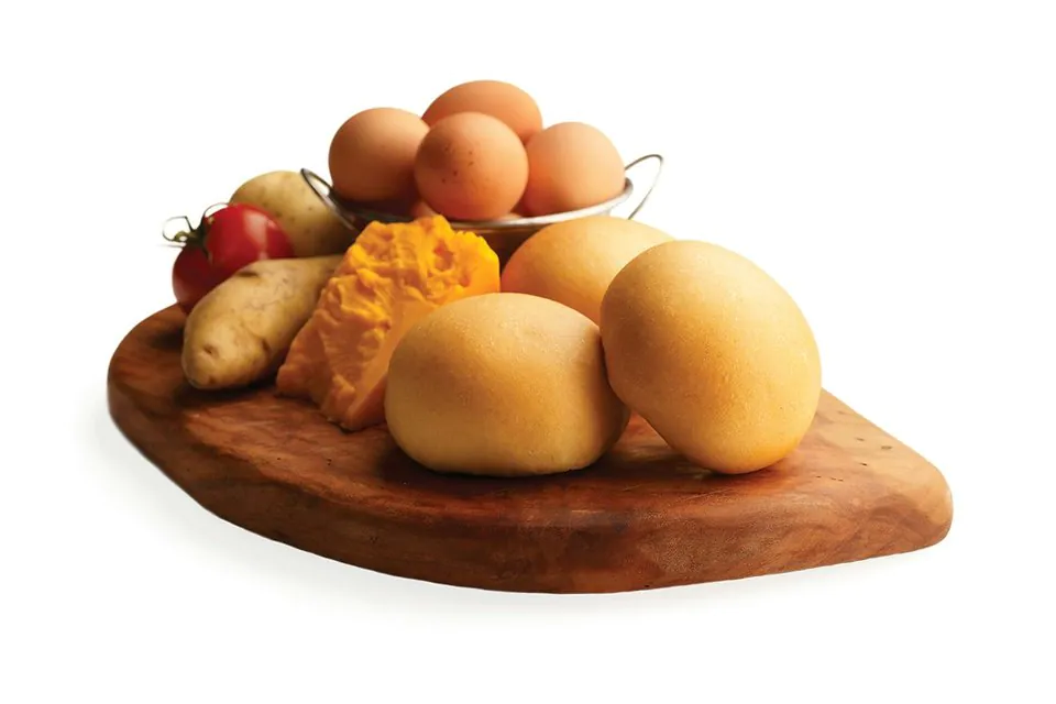 Four golden Potato, Egg and Cheese kolaches on a wooden board with a bowl of brown eggs, a wedge of cheddar and a potato behind them