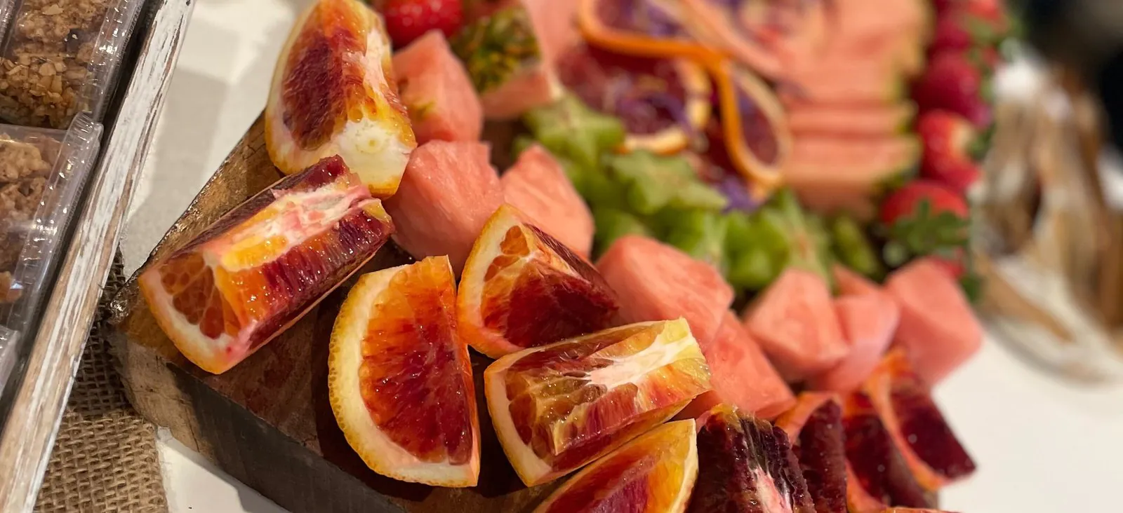 Blood orange wedges and cubes of watermelon on a wooden fruit board, with strawberries and grapes behind