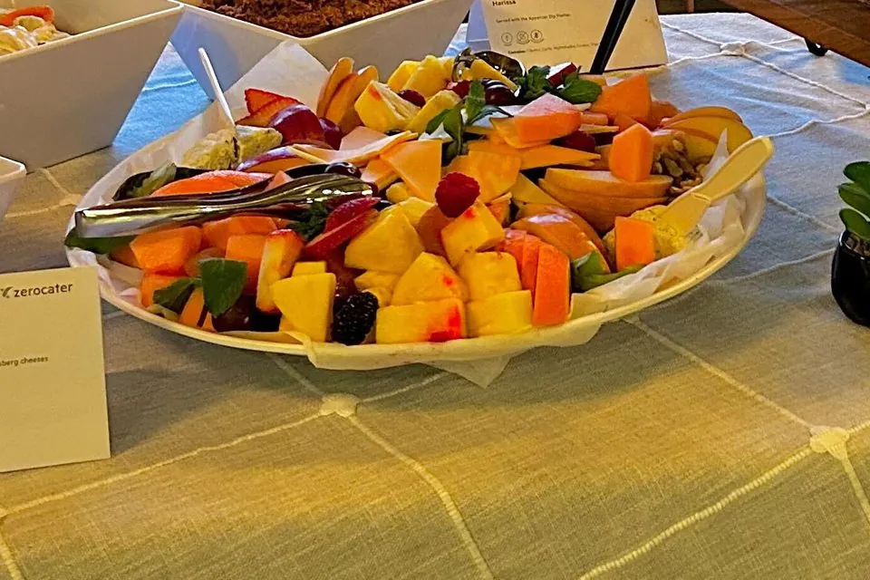 Round catering platter of cut pineapple, melon, apple slices, berries and wedges of hard cheese on a buffet table, with a Zerocater table card beside it