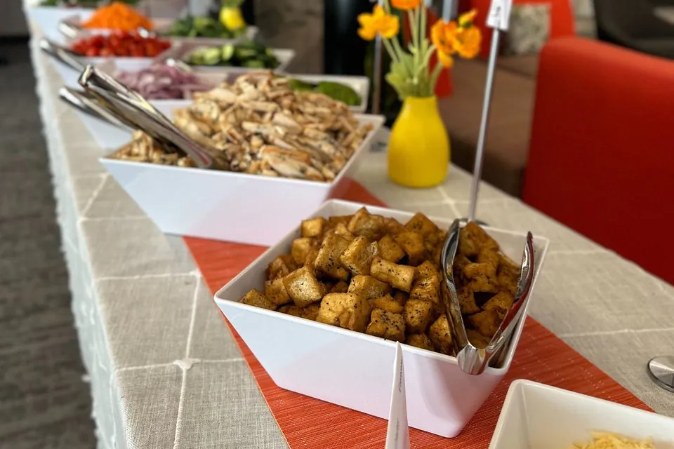 Office salad bar line in white square bowls with plain sliced grilled chicken, red onion, cucumbers, carrots and seasoned croutons