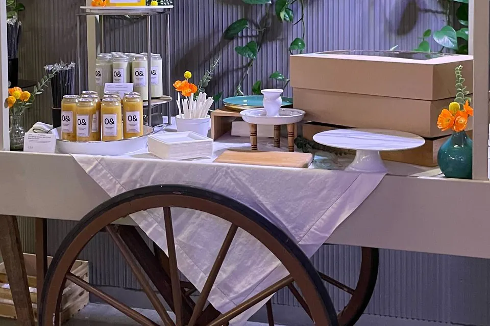 Glass bottles of mango and pale juice drinks on a wooden cart with a white cloth, flowers and a cake stand