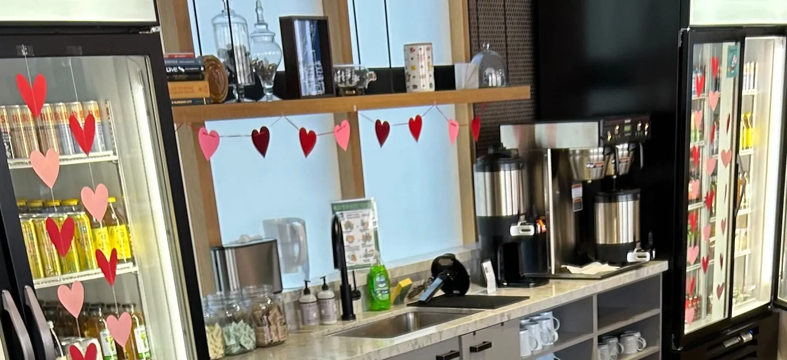 Office pantry with two glass-door drink fridges, a batch coffee brewer with two insulated servers, jars of tea and a shelf of white mugs