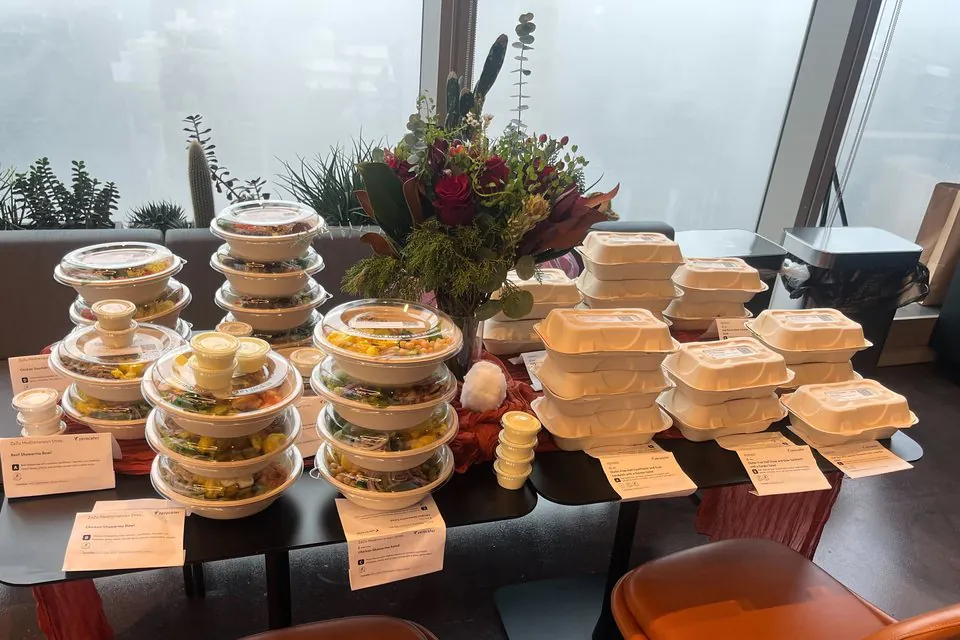 Stacks of clear-lidded salad bowls and white clamshell boxed meals with dish cards on an office table beside a floral centerpiece