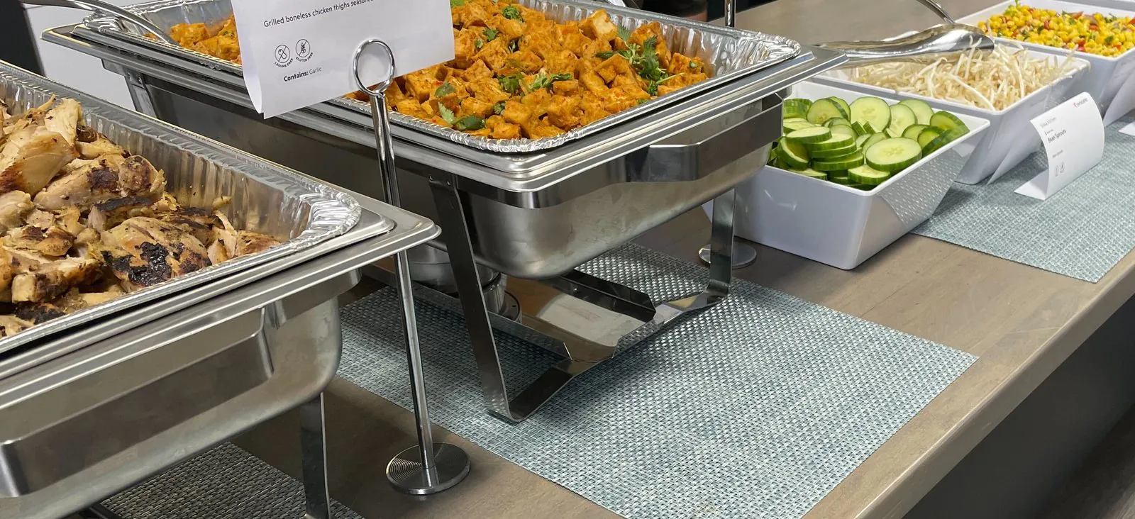 Two chafing trays of hot dishes with a grilled lemongrass chicken card beside white bowls of sliced cucumber and bean sprouts on an office buffet