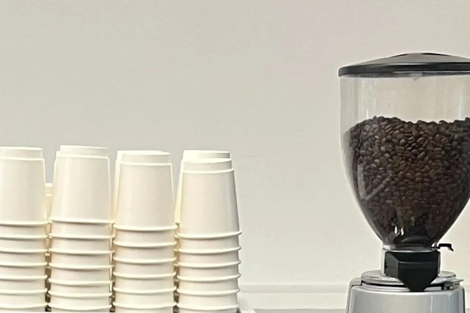 Clear grinder hopper full of whole roasted coffee beans beside four stacks of white paper cups against a white wall