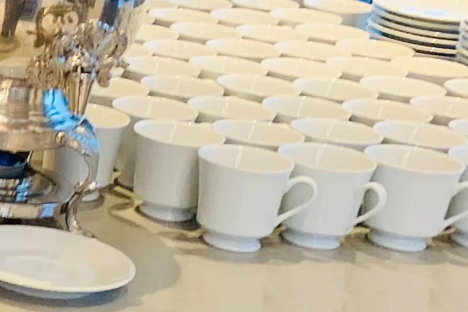 Rows of white porcelain coffee cups on a counter beside the silver base of a coffee urn