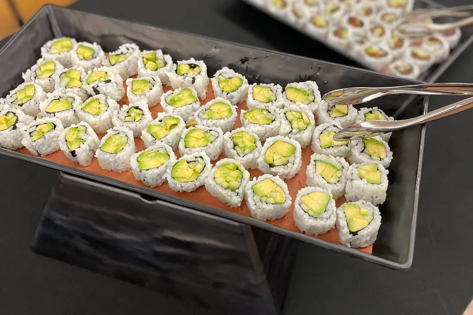 Black rectangular tray of avocado and cucumber maki rolls with serving tongs, a second tray of rolls behind it