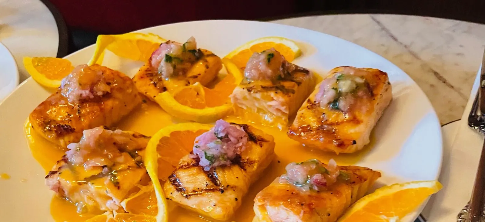 Grilled salmon fillets topped with a shallot and herb relish on a white platter with orange slices