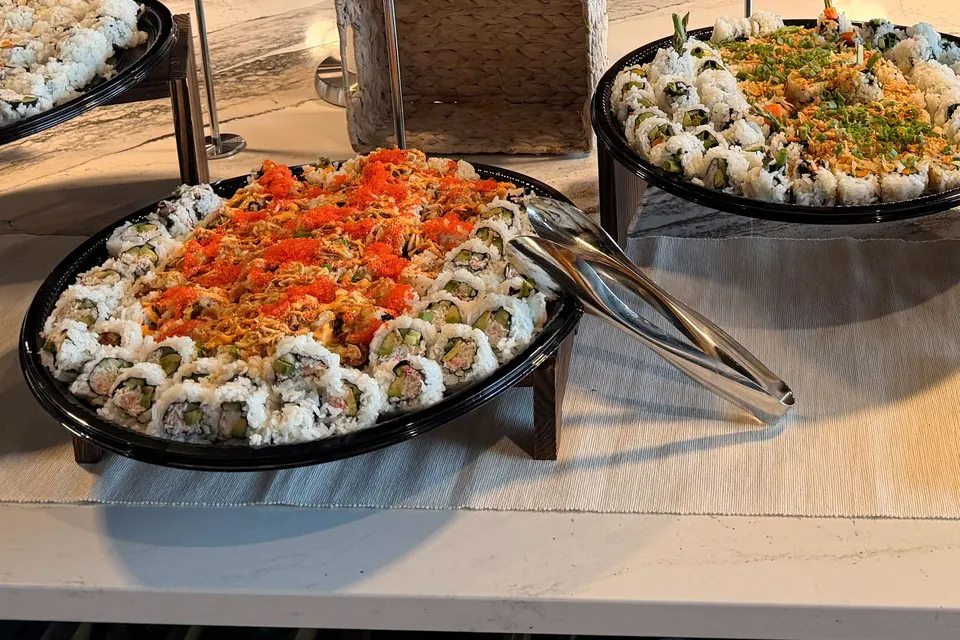 Two round black platters of sushi rolls on wooden risers on an office buffet counter, one topped with orange fish roe, with serving tongs