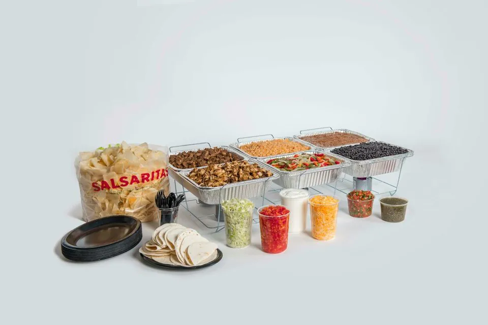 A Salsarita's Fajita Bar: foil trays of grilled steak and grilled chicken, Mexican rice, sauteed fajita vegetables, refried and black beans on wire racks, with a stack of flour tortillas, a bag of chips, black plates and tubs of lettuce, tomato, sour cream, cheese, pico de gallo and salsa verde