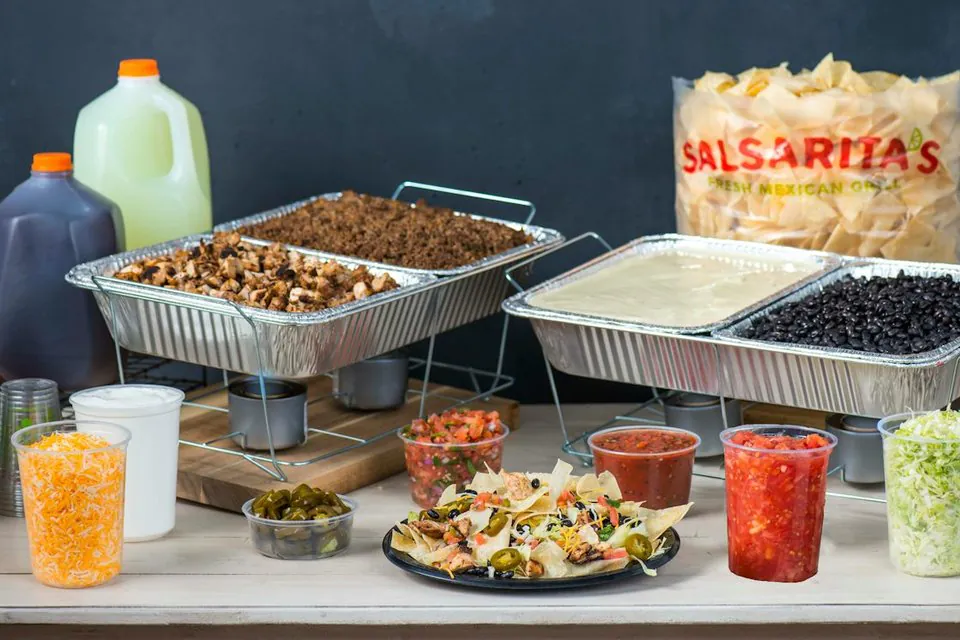 A Salsarita's Nacho Bar: a foil tray of grilled chicken and seasoned ground beef beside trays of queso and black beans on wire racks, gallon jugs of tea and lemonade, a bag of Salsarita's chips, and tubs of cheese, sour cream, jalapenos, pico de gallo and salsas around a plate of loaded nachos