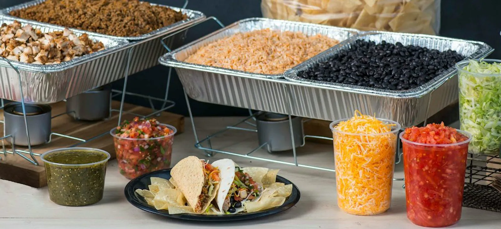 A Salsarita's Taco Bar set up for an office: foil trays of grilled chicken and seasoned ground beef, Mexican rice and black beans on wire chafing racks, with tubs of shredded cheese, pico de gallo and salsa verde and a plate of two tacos