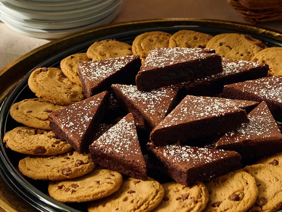 A Sonny's BBQ dessert platter of chocolate chip cookies ringed around powdered-sugar brownie triangles on a black tray, with a stack of plates behind