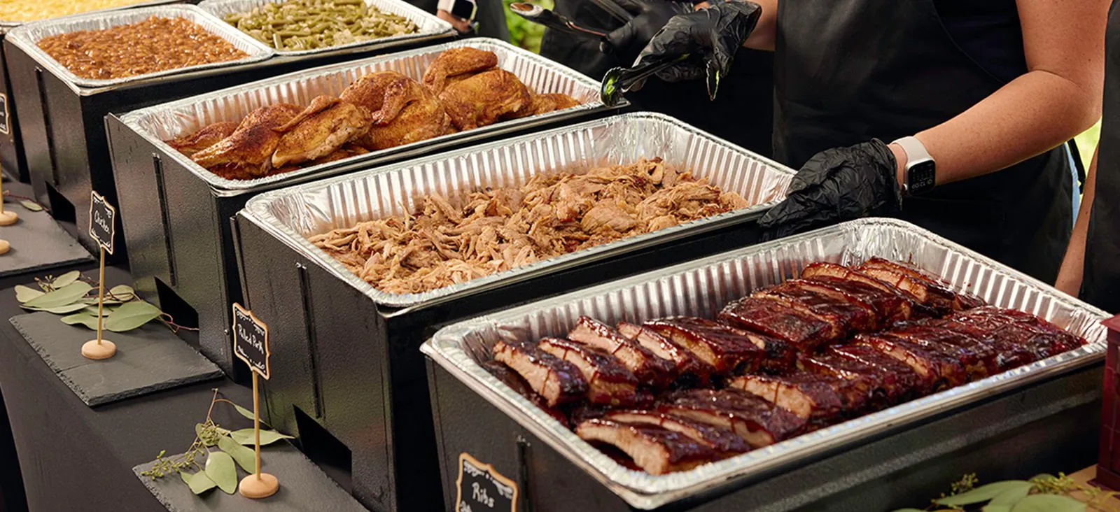 A Sonny's BBQ catering line of foil pans in black chafers: BBQ beans, green beans, BBQ chicken, pulled pork and a pan of sauced ribs, with gloved hands serving behind