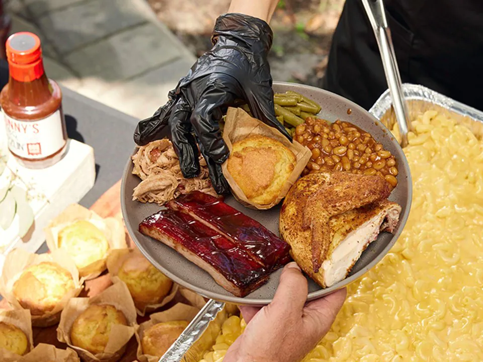 A gloved hand holding a Sonny's BBQ plate of pulled pork, a sauced rib, BBQ chicken, a cornbread muffin, BBQ beans and green beans over a foil pan of mac and cheese, with a Sonny's sauce bottle at the edge