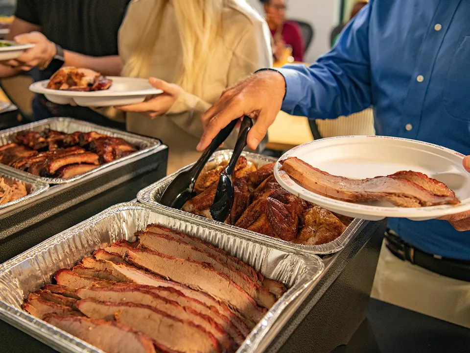 Sliced Sonny's BBQ brisket in a foil pan on an office catering line, with a hand lifting a slice onto a white plate with tongs and a pan of BBQ chicken behind