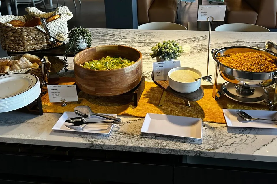 Basket of dinner rolls, a wooden bowl of salad, a bowl of dressing and a chafing dish of corn on a marble office counter