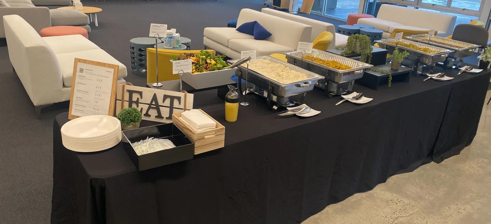 Long office buffet on a black tablecloth with chafing trays, a salad bowl, a stack of plates and a wooden EAT sign in an open lounge