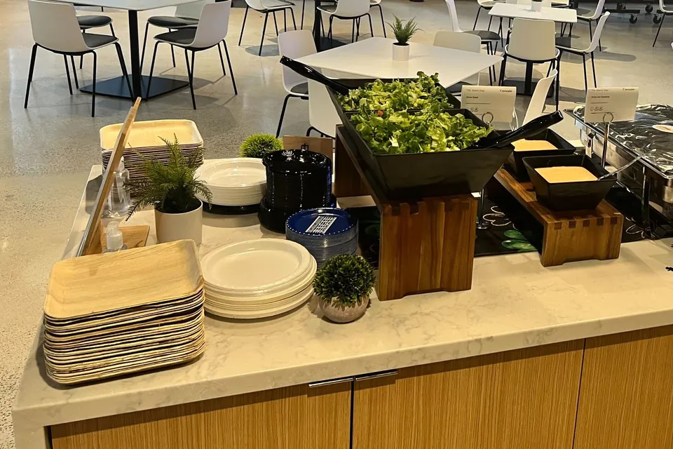 Black bowl of mixed greens on a wooden riser beside stacks of palm-leaf and white plates at an office cafe buffet
