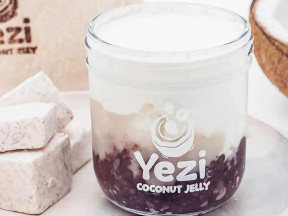 A glass jar of Yezi Taro Purple Rice Coconut, with purple rice and taro under white coconut milk jelly, beside blocks of taro