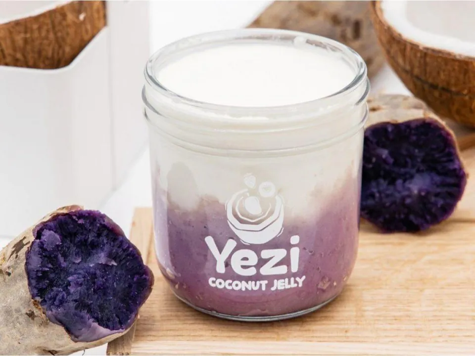 A glass jar of Yezi Ube Taro Coconut, with purple ube and taro under white coconut milk jelly, beside a cut purple sweet potato