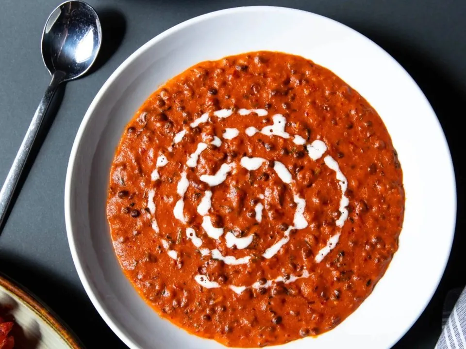 A white bowl of black lentil dal with a swirl on top and a spoon beside it