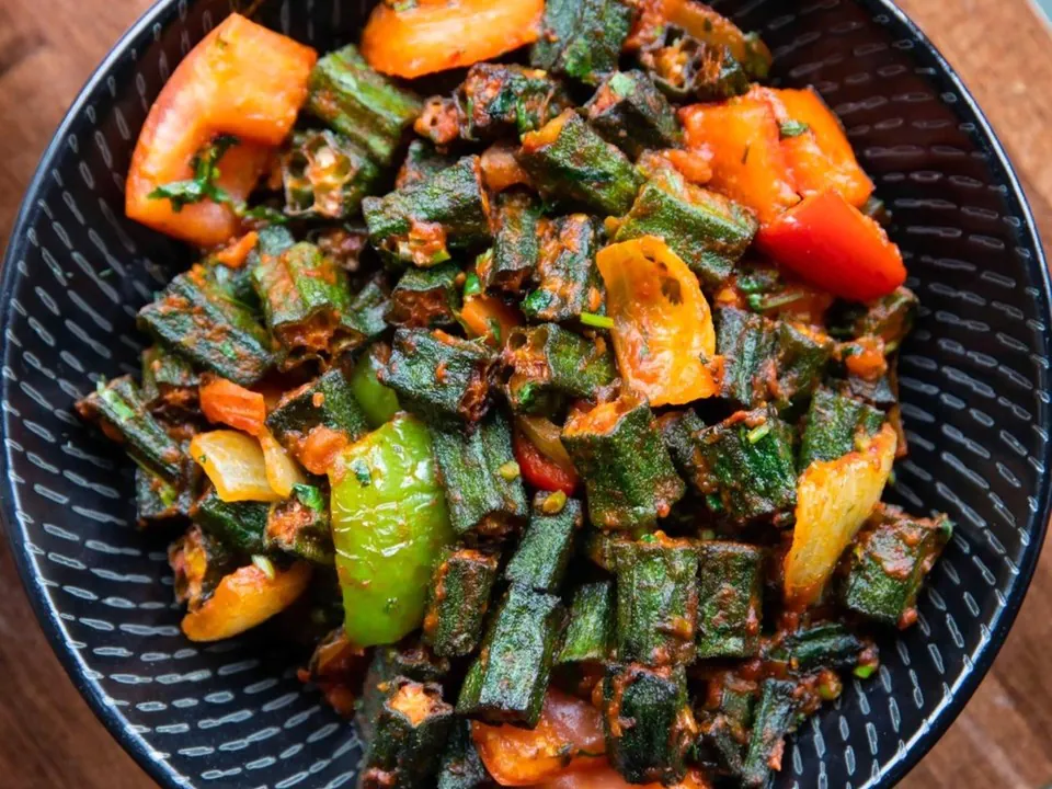 A dark bowl of okra cooked with tomato, onion and green and orange peppers