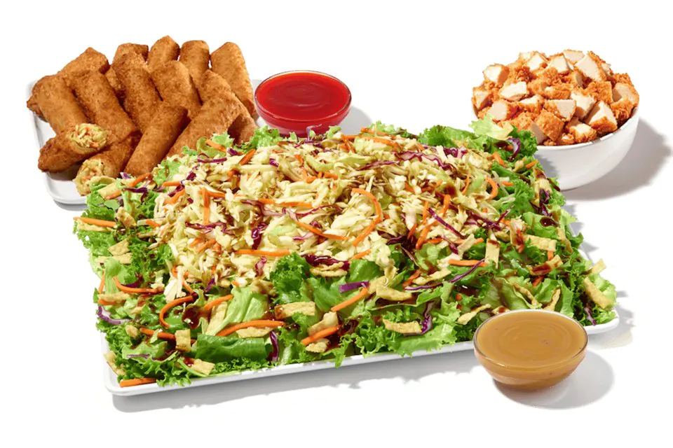 A Zaxby's Asian Zensation Zalad Platter of greens, slaw and wonton strips beside a tray of veggie egg rolls, a bowl of fried chicken pieces and cups of sauce and dressing